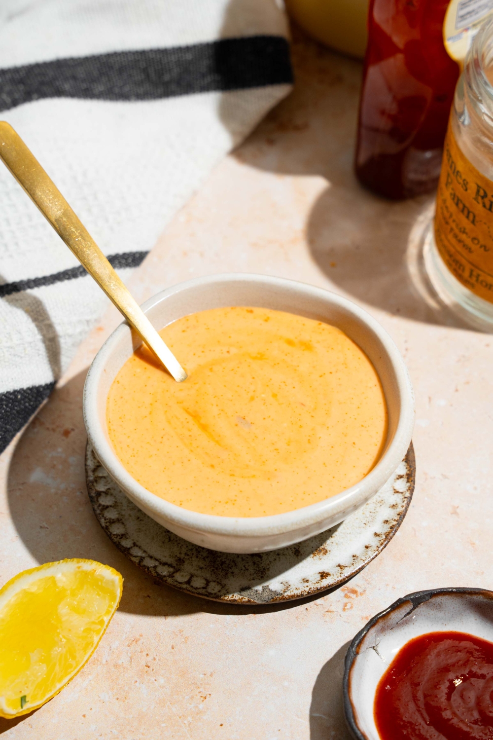 A bowl of honey sriracha sauce with a spoon in the bowl. The bowl is on a tan counter with a jar of honey and bowl of sriracha.
