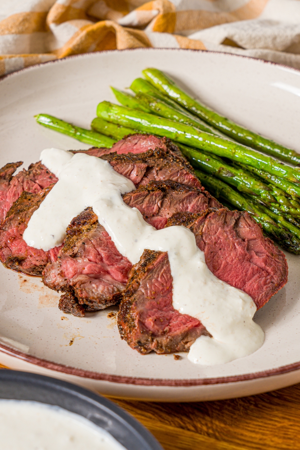 A white plate with sliced steak and asparagus, with horseradish sauce drizzled over the steak. The plate is on a wooden counter with a yellow checkered napkin.
