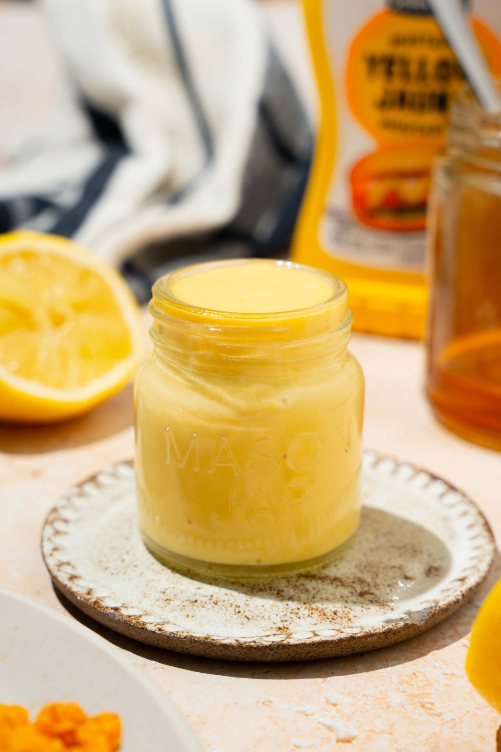 A jar of honey mustard sauce on a saucer. The jar is on a tan counter with a lemon, jar of honey, and mustard.
