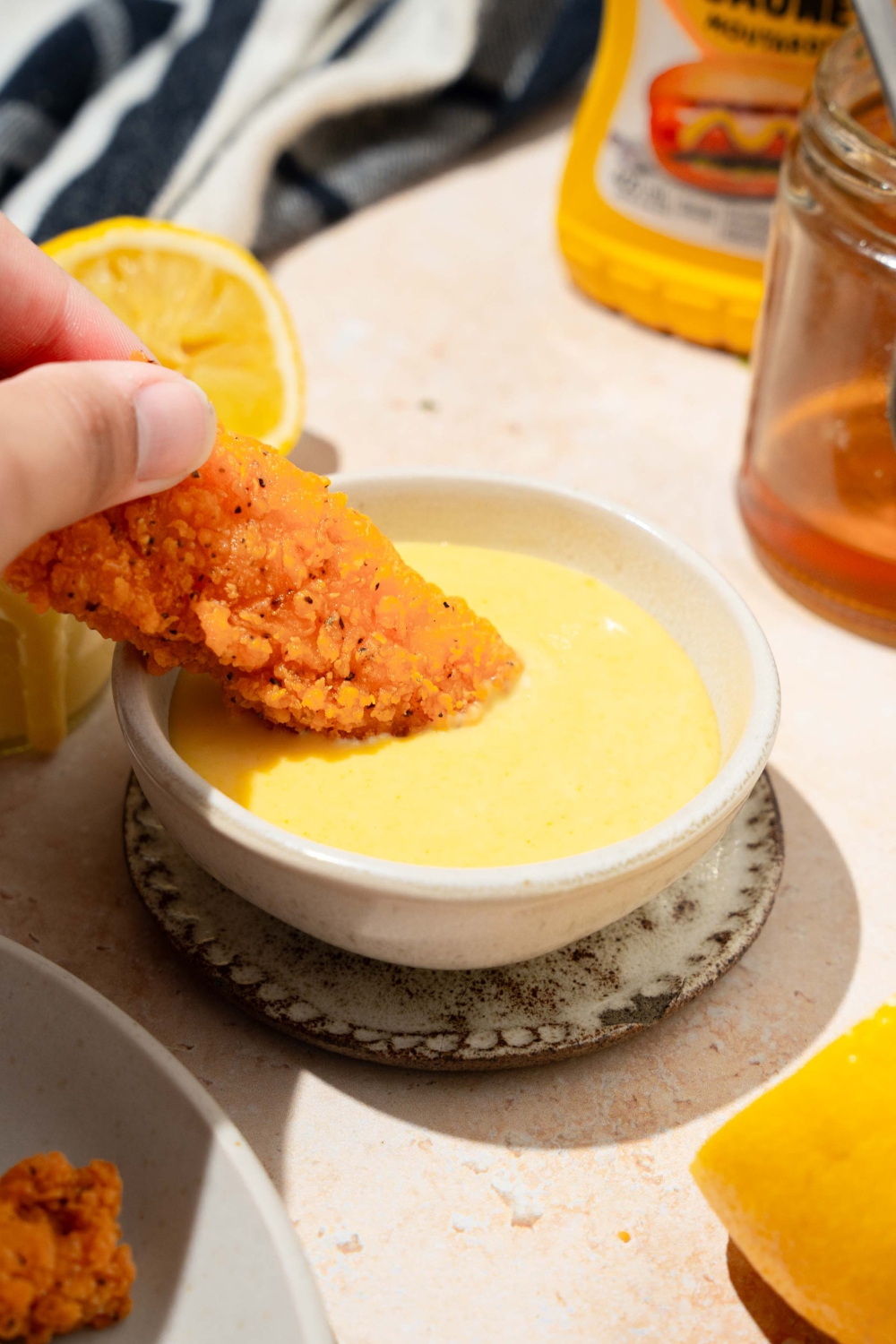 A bowl of honey mustard sauce with a chicken tender dipped in the sauce. The bowl is on a saucer on a tan counter with a lemon and jar of honey.