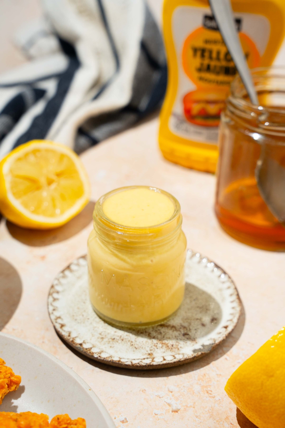 A jar of honey mustard sauce on a saucer. The jar is on a tan counter with a lemon, jar of honey, and mustard.