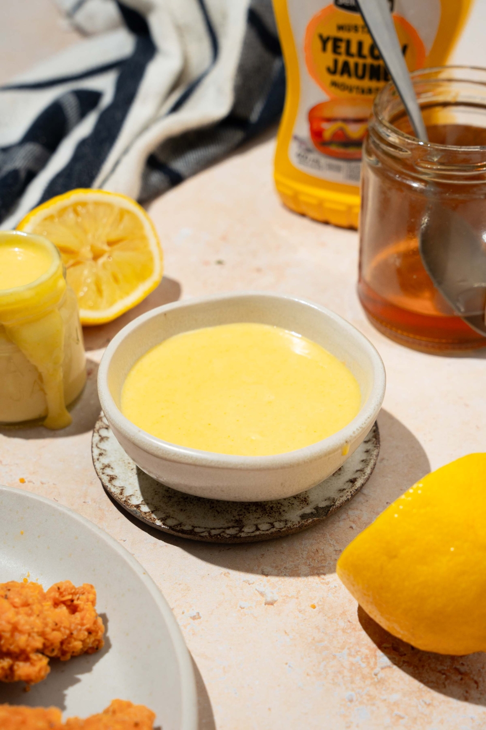 A bowl of honey mustard sauce on a saucer. The bowl is on a tan counter with a lemon, a plate of chicken tenders, and a jar of honey.