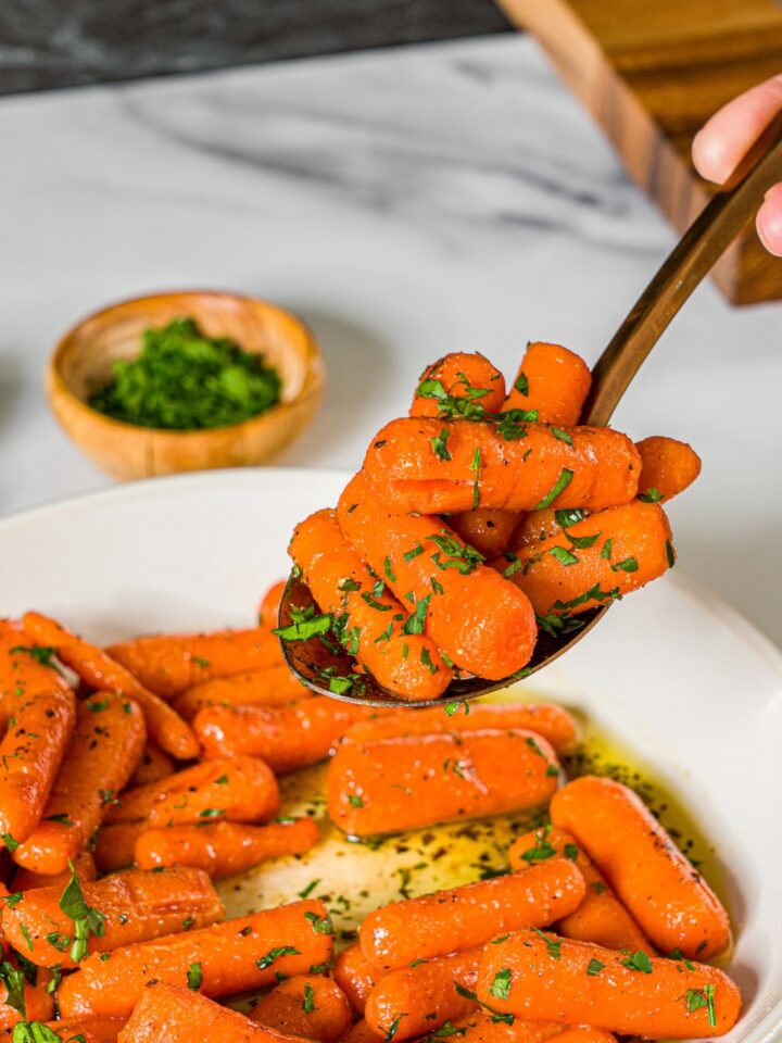 A spoon with a bite of honey roasted baby carrots garnished with fresh parsley. There is a bowl of carrots on a marble counter and small bowl of parsley.