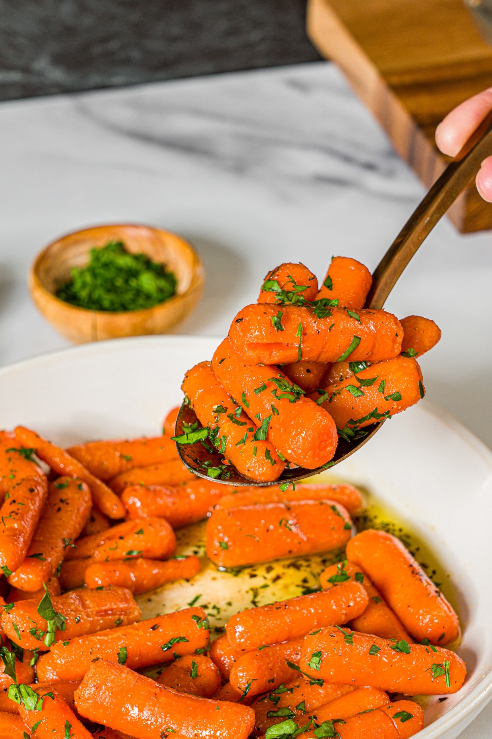 A spoon with a bite of honey roasted baby carrots garnished with fresh parsley. There is a bowl of carrots on a marble counter and small bowl of parsley.
