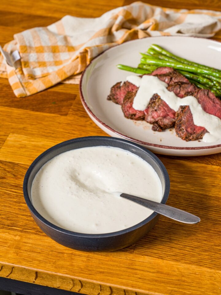 A white plate with sliced steak and asparagus, with horseradish sauce drizzled over the steak. The plate is on a wooden counter with a yellow checkered napkin and a bowl of horseradish sauce.