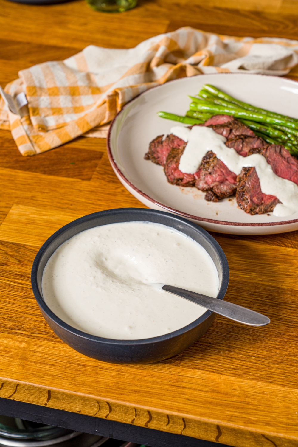 A white plate with sliced steak and asparagus, with horseradish sauce drizzled over the steak. The plate is on a wooden counter with a yellow checkered napkin and a bowl of horseradish sauce.