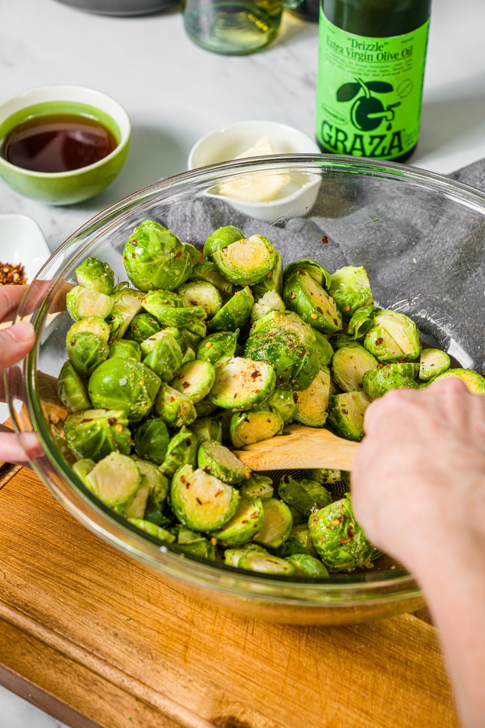 A large glass bowl with a spoon mixing trimmed brussels sprouts with oil and seasonings. The bowl is on a wooden board on a marble counter with bowls of ingredients.