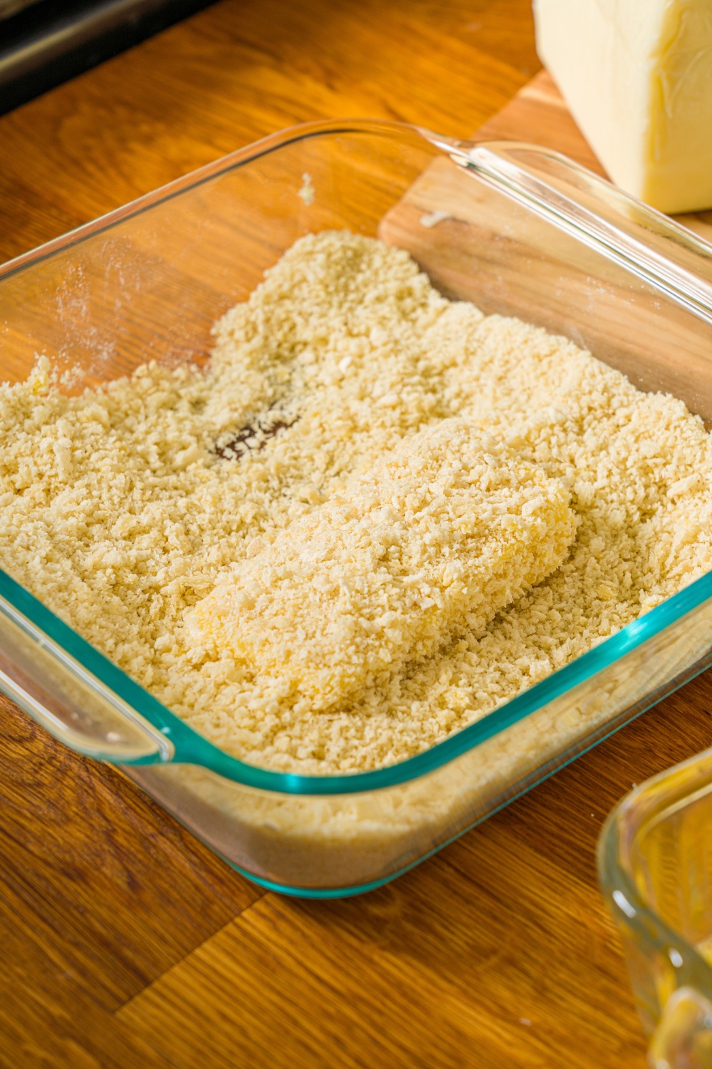 A glass baking dish with mozzarella being breaded in breadcrumbs. The dish is on a wooden counter.