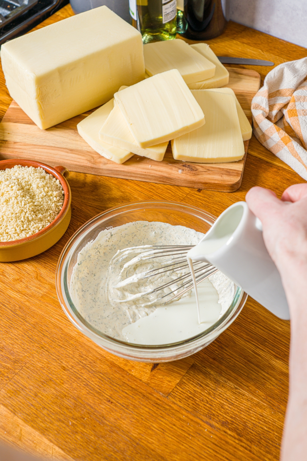 A glass bowl with a whisk and ranch mixture with buttermilk being poured into the mixture. The bowl is on a wooden counter with a board of mozzarella cheese.