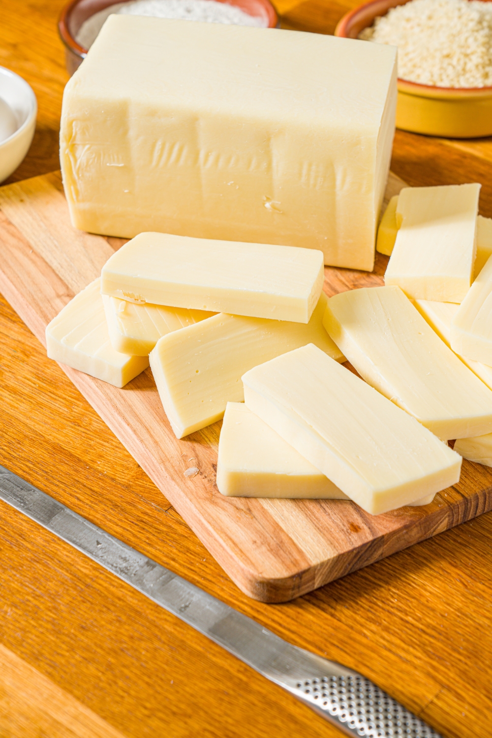 A wooden board with a block of mozzarella cheese sliced into sticks. The board is on a wooden counter.
