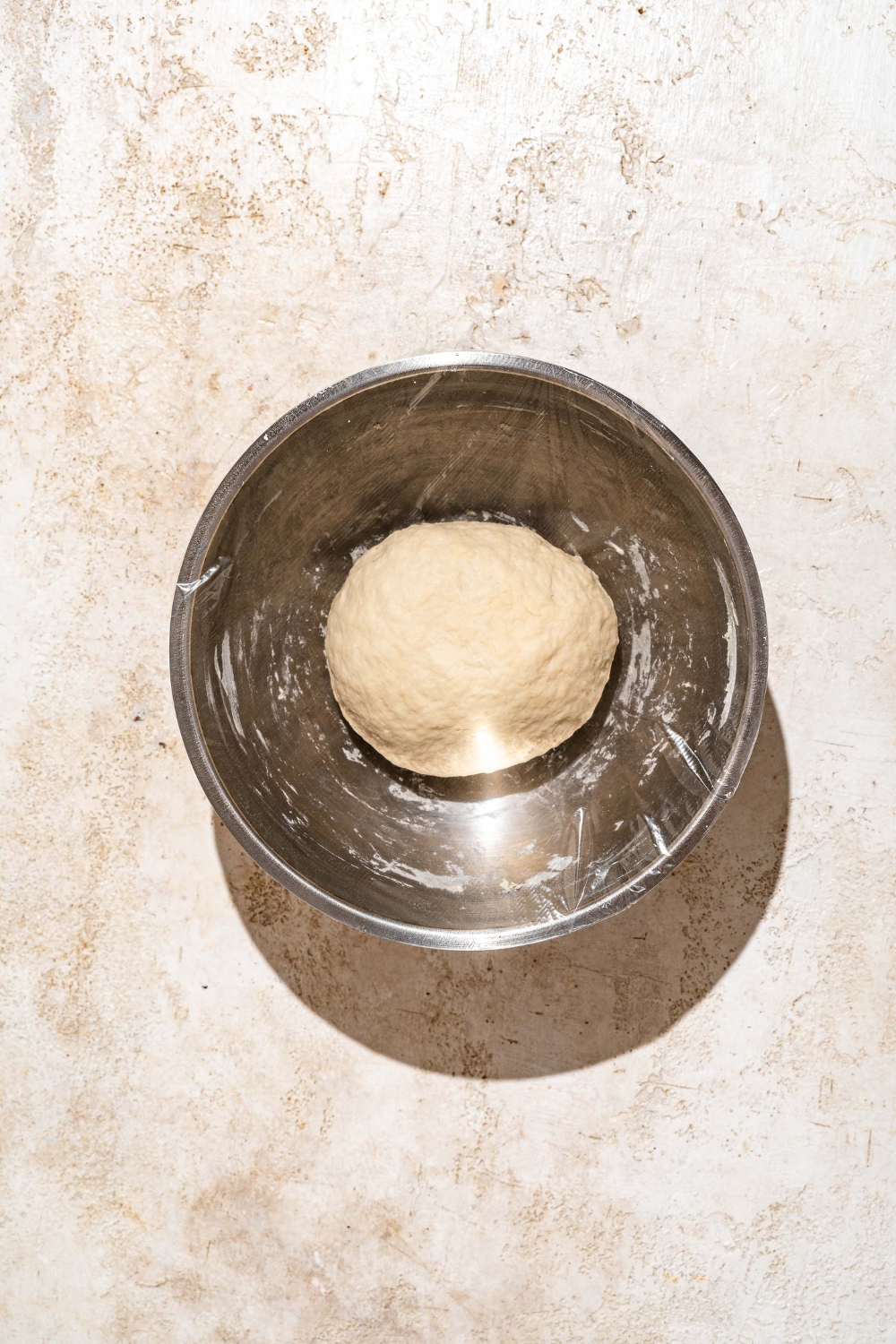 A bowl with a large ball of Indian fry bread dough wrapped in plastic wrap. The bowl is on a tan counter.