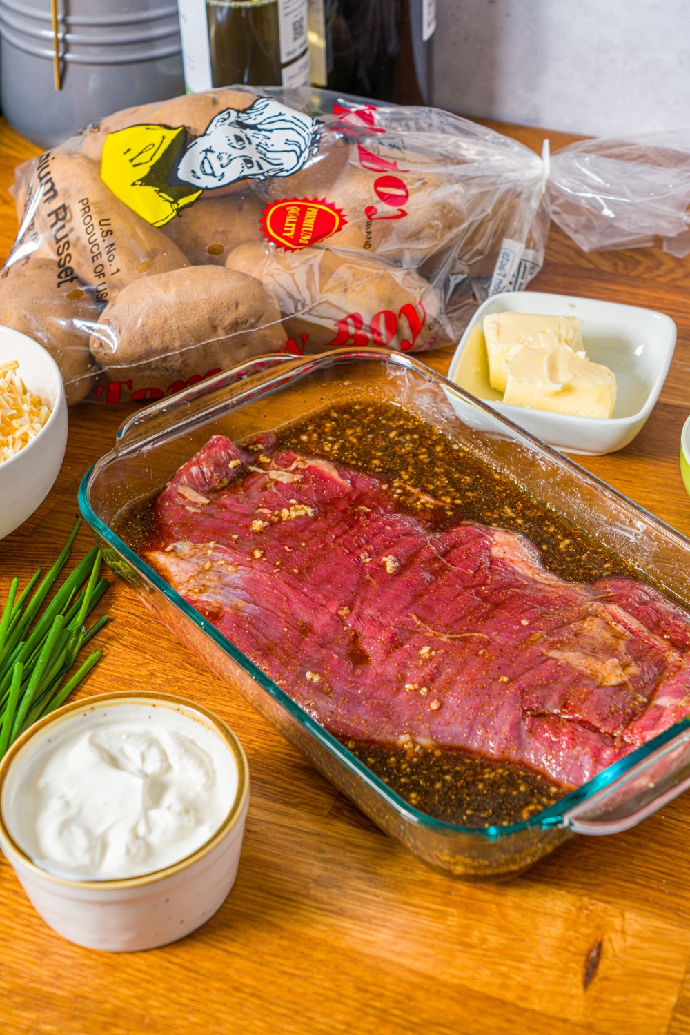 A whole steak marinating in a glass baking dish filled with marinade. The dish is on a wooden counter with ingredients to make steak loaded potato skins.