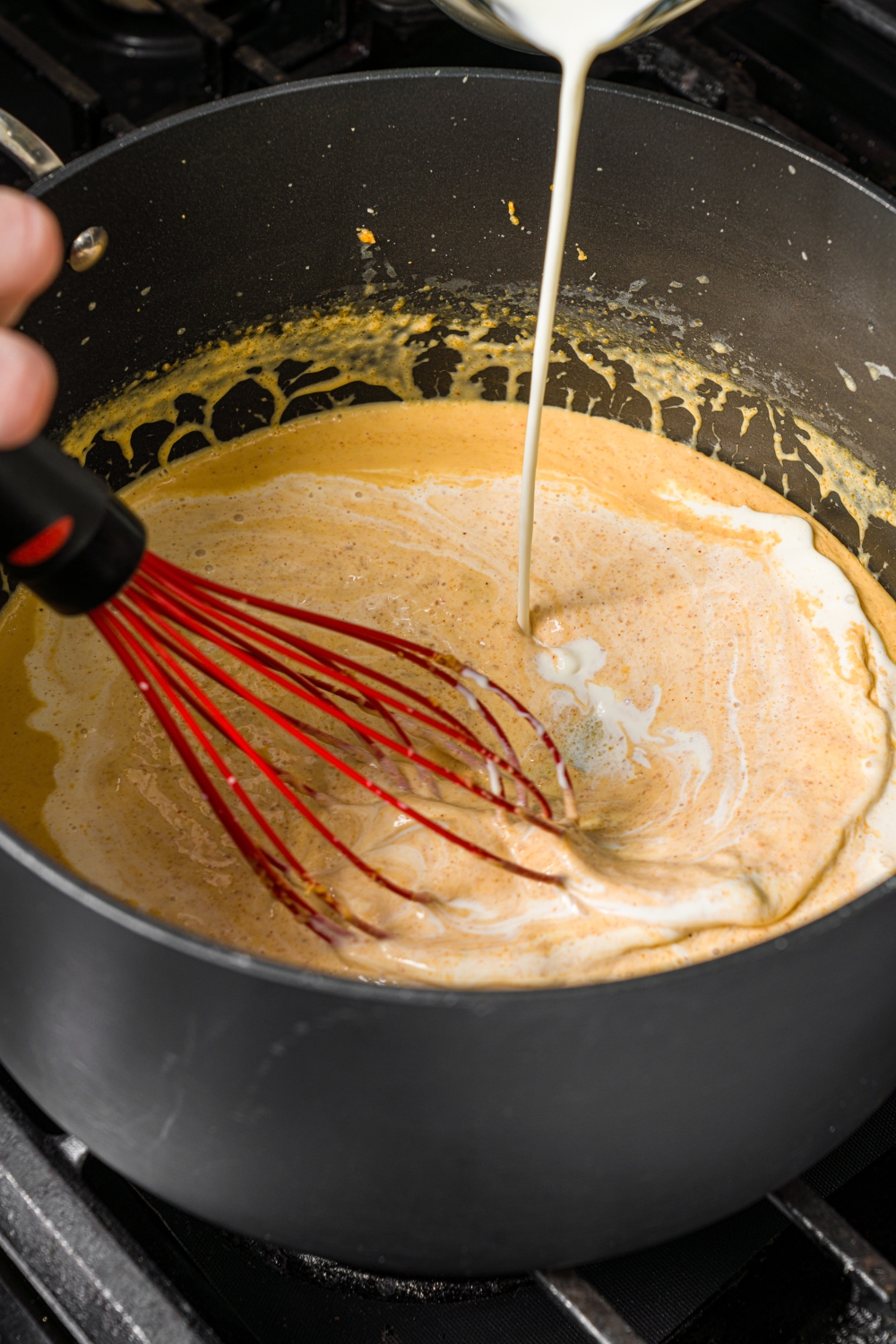A large pot with a whisk mixing ingredients to make macaroni and cheese sauce while cream is being added to the mixture.