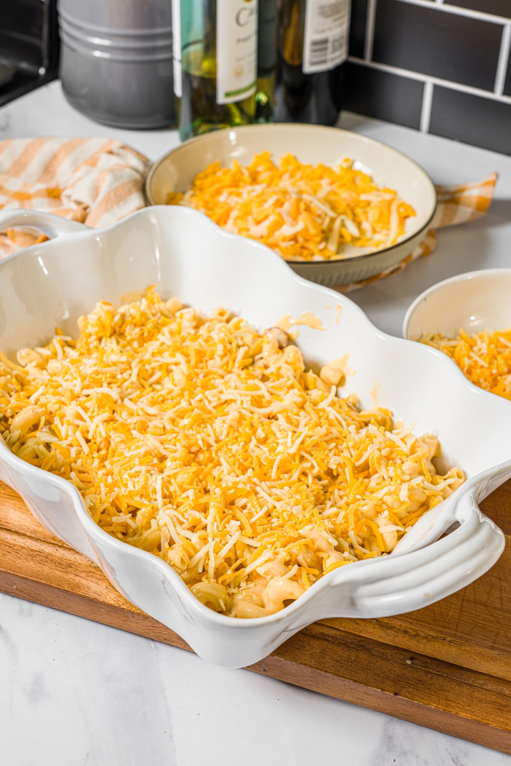 A baking dish with bacon macaroni and cheese topped with shredded cheese. The dish is on a wooden counter with bowls of cheeses.
