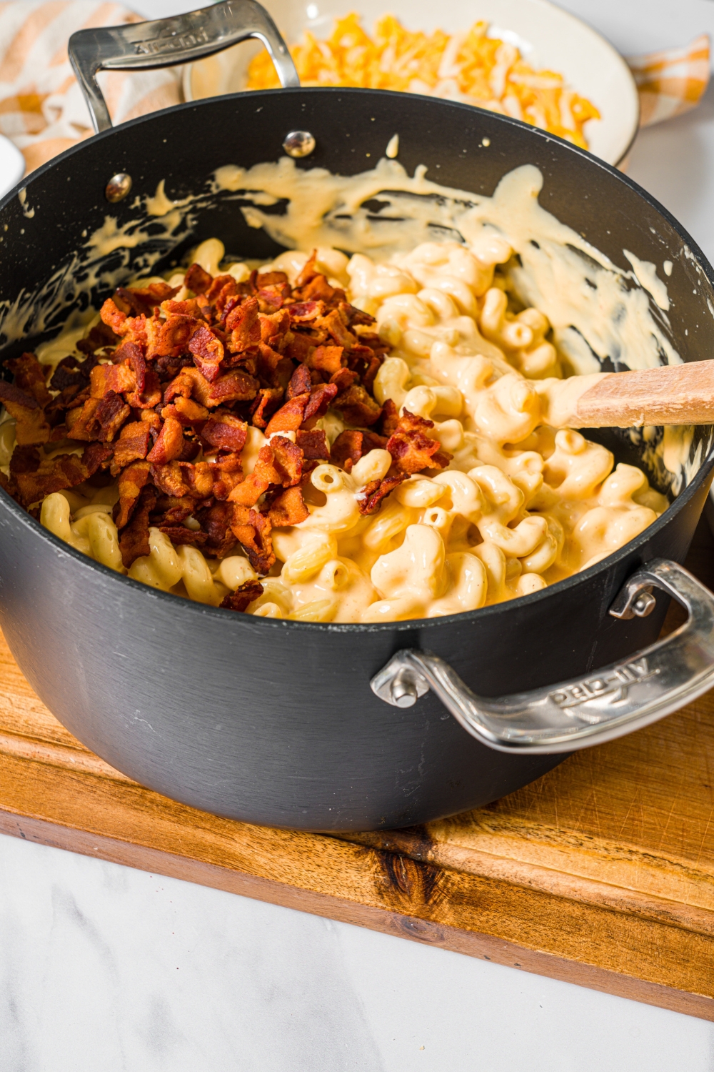A large pot with cavatappi pasta and crispy bacon being mixed with macaroni and cheese sauce. The pot is on a wooden counter.