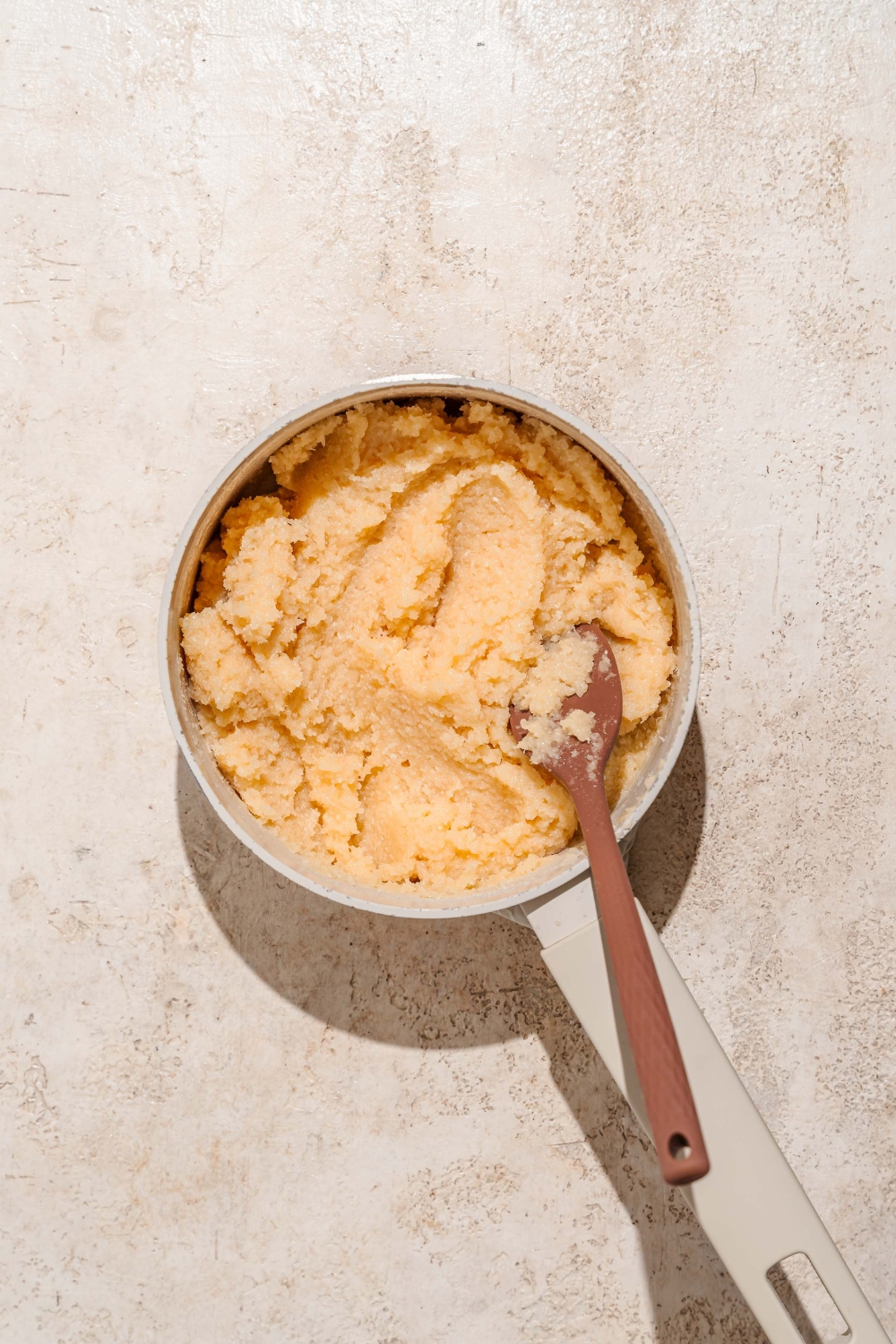 A saucepan with a bala baiana dough mixture. A wooden spoon is stirring the dough. The saucepan is on a tan counter.