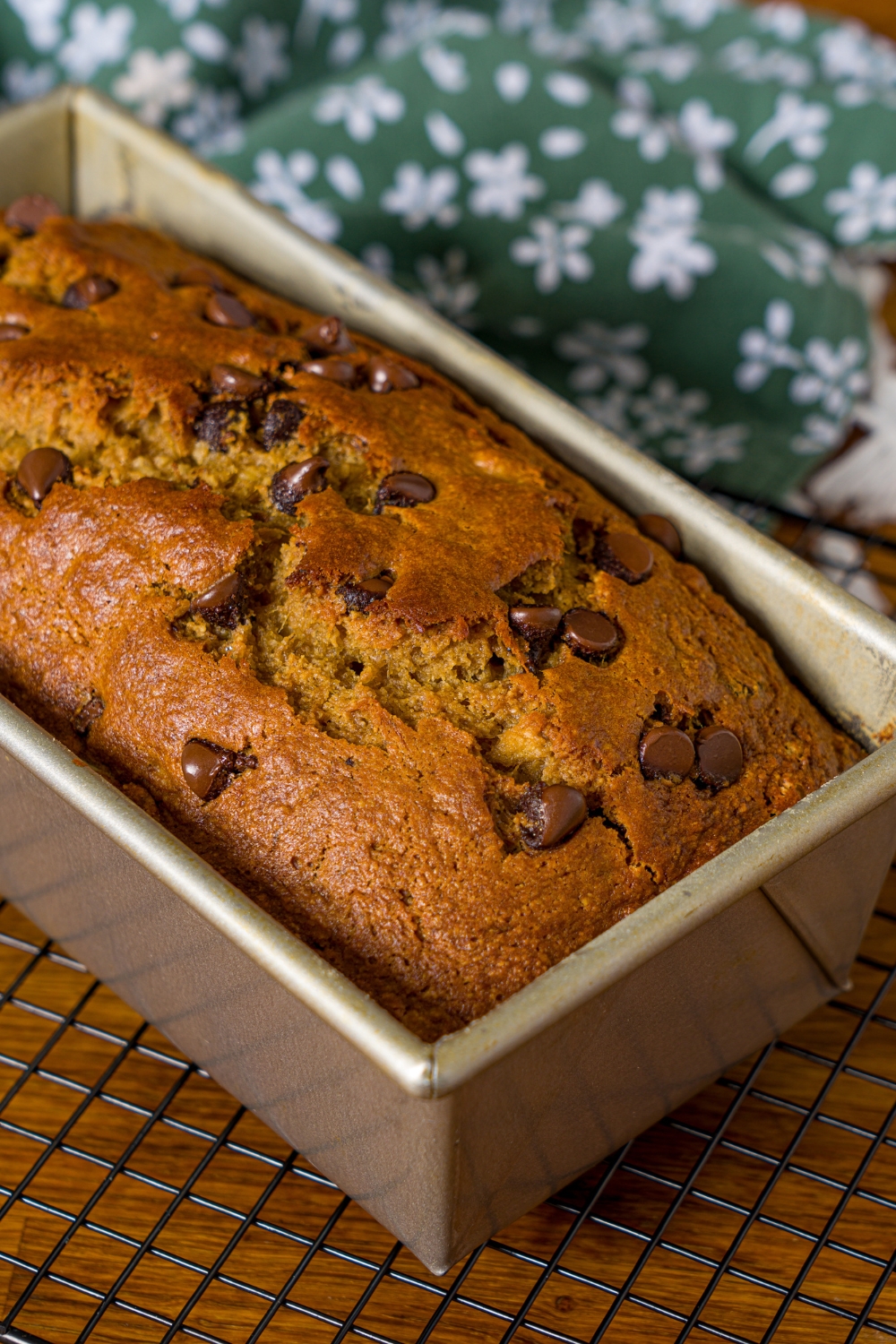A loaf pan with baked chocolate chip banana bread with olive oil. The pan is on a wire rack on a wooden counter with a green floral napkin.