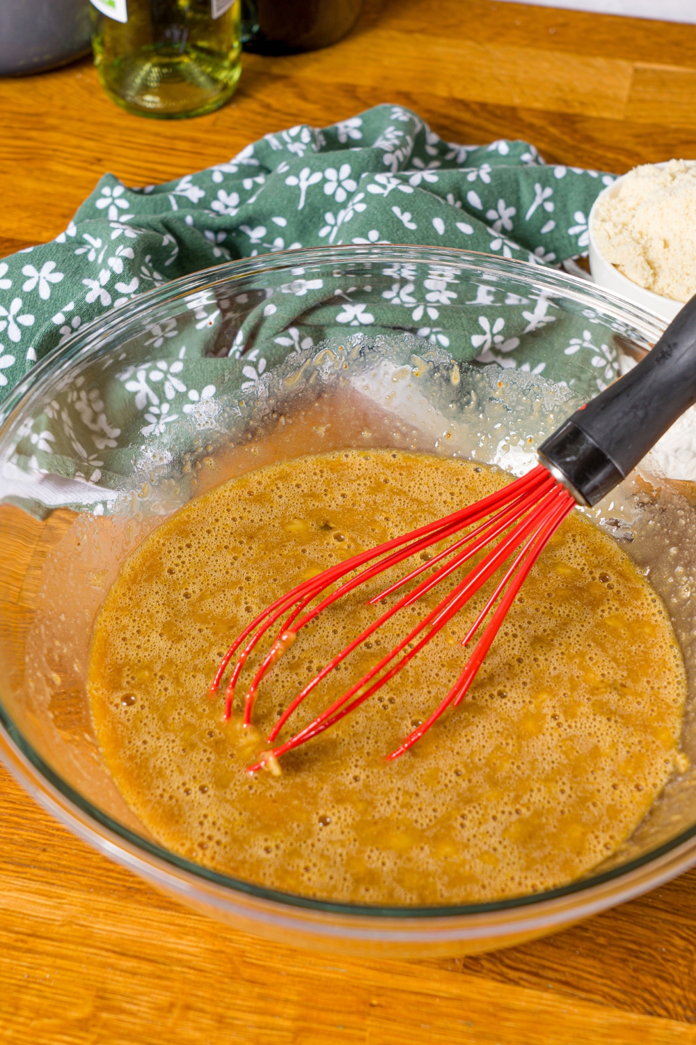 A large bowl with a whisk mixing wet ingredients to make banana bread with olive oil. The bowl is on a wooden counter with a green floral napkin.