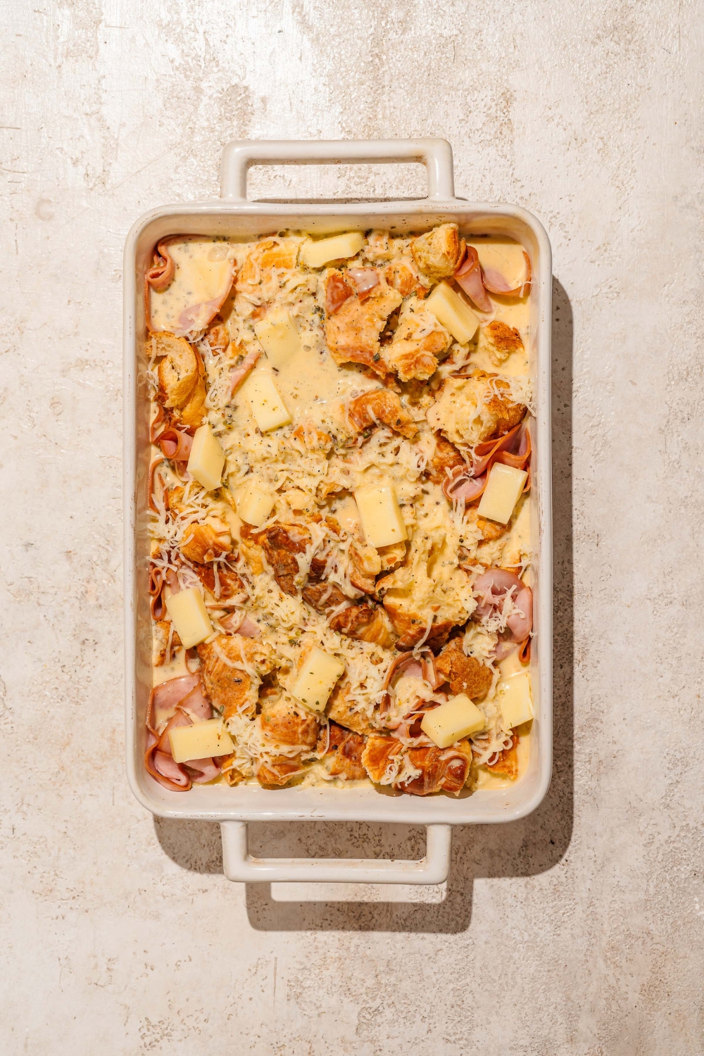 A baking dish with croissant, ham, and cheese breakfast casserole mixture soaking in custard. The dish is on a tan counter.
