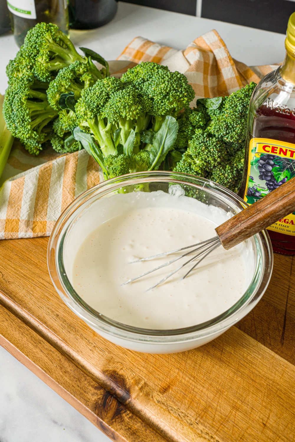 A glass bowl with a whisk mixing greek yogurt dressing. The bowl is on a wooden board with broccoli and a yellow checkered napkin.