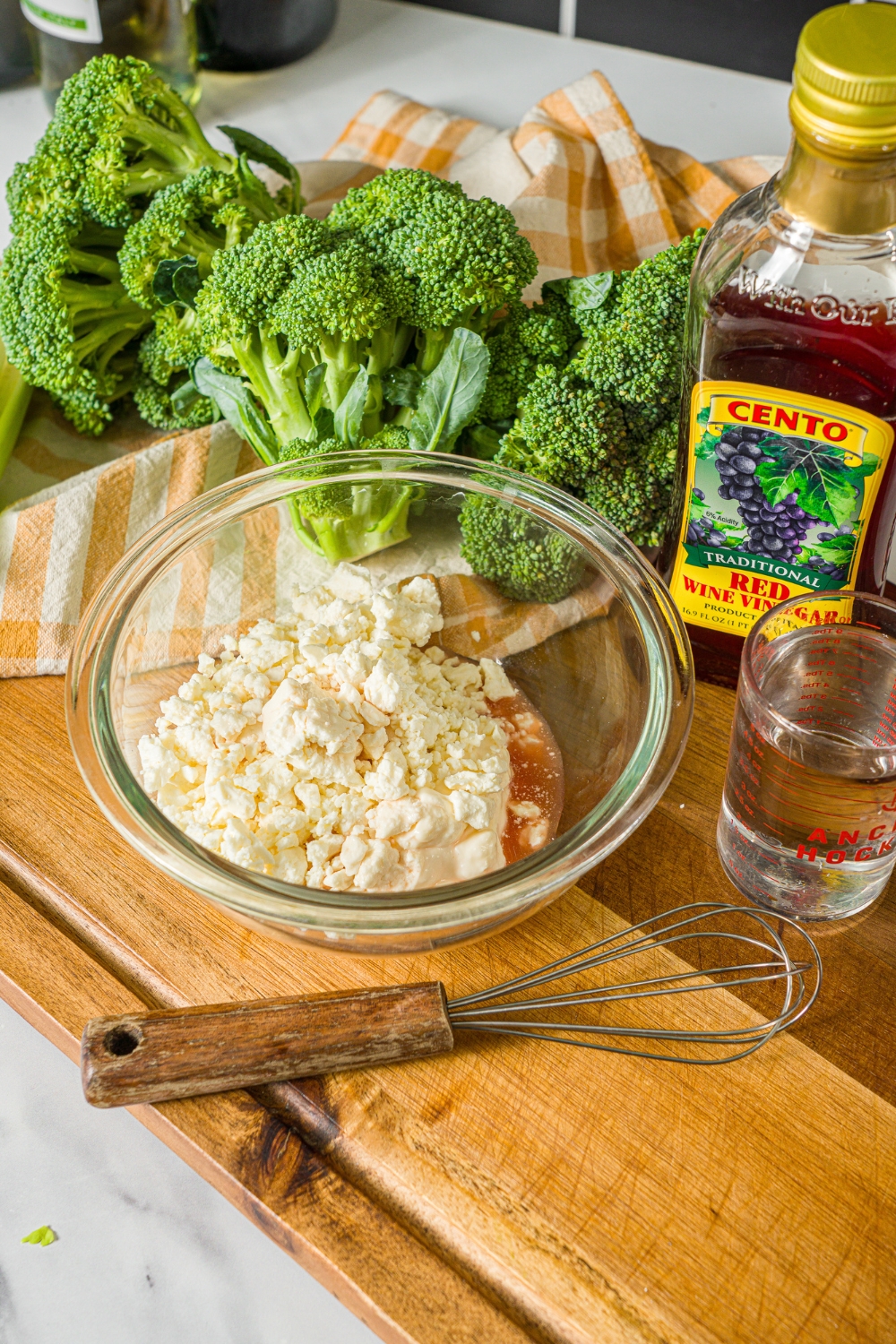 A glass bowl with ingredients to make greek yogurt dressing including greek yogurt, feta cheese, and red wine vinegar. The bowl is on a wooden board with a whisk and broccoli.
