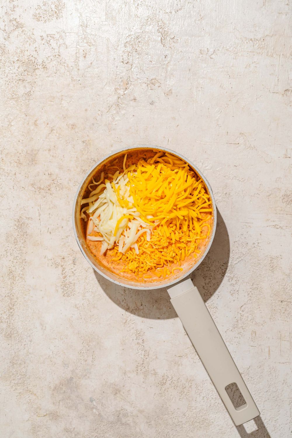 A saucepan with a sauce base topped with shredded cheeses. The saucepan is on a tan counter.