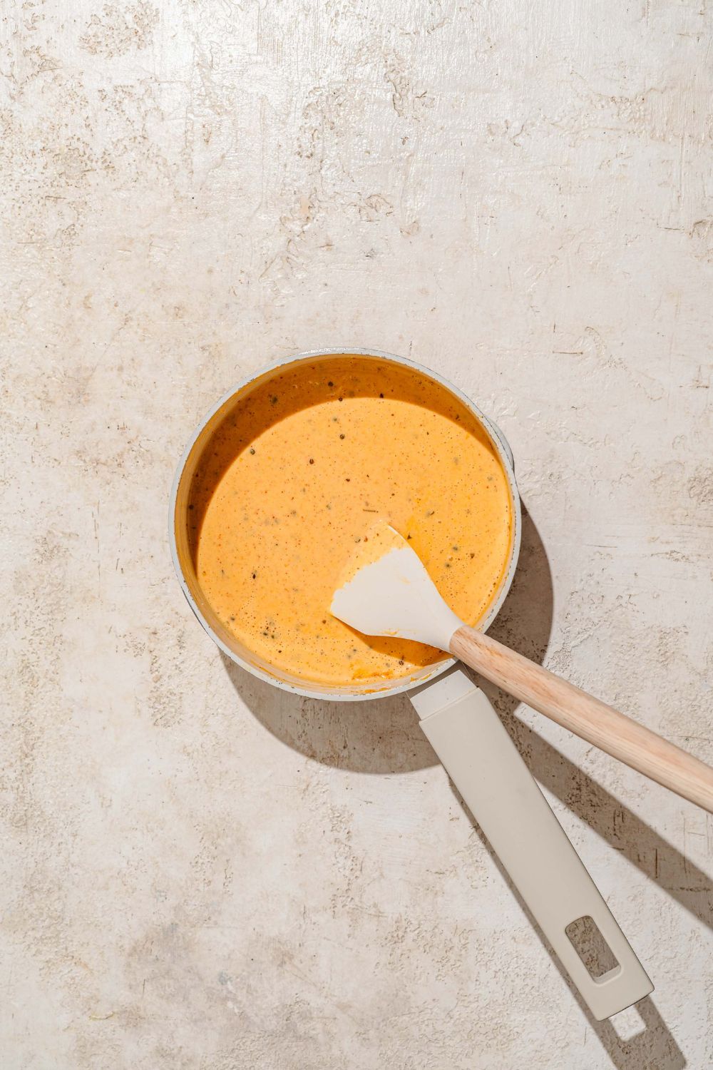 A saucepan with a spatula mixing a seasoned cheddar cheese sauce. The saucepan is on a tan counter.