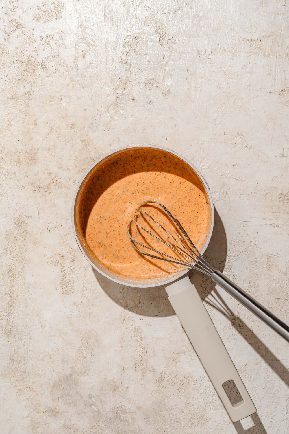 A saucepan with a seasoned cheddar cheese sauce base with a whisk. The saucepan is on a tan counter.