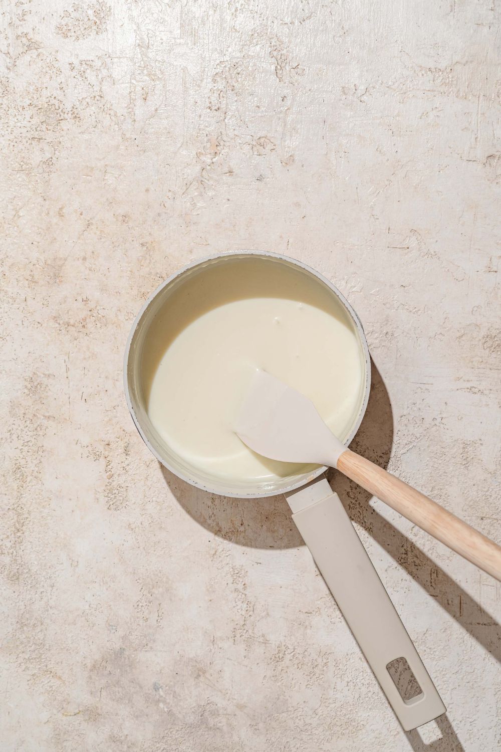 A saucepan with a spatula mixing a roux and milk base. The saucepan is on a tan counter.