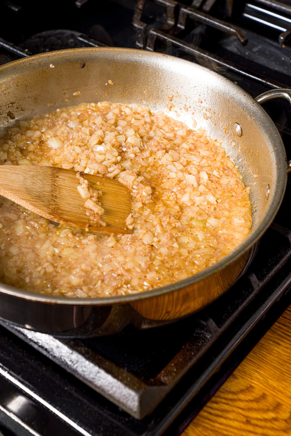A skillet with garlic, onion, and shallots sautéing in oil and deglazed with white wine. The skillet is on a stove with a spoon stirring the mixture.