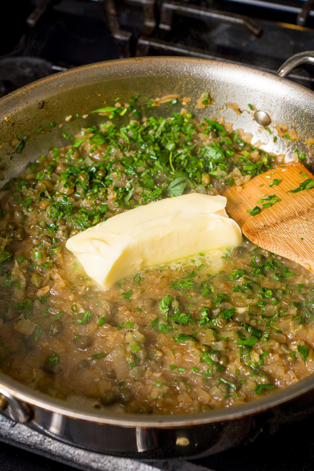 A skillet with chicken piccata sauce with butter melting in the sauce. The skillet is on a stove with a spoon stirring the mixture.