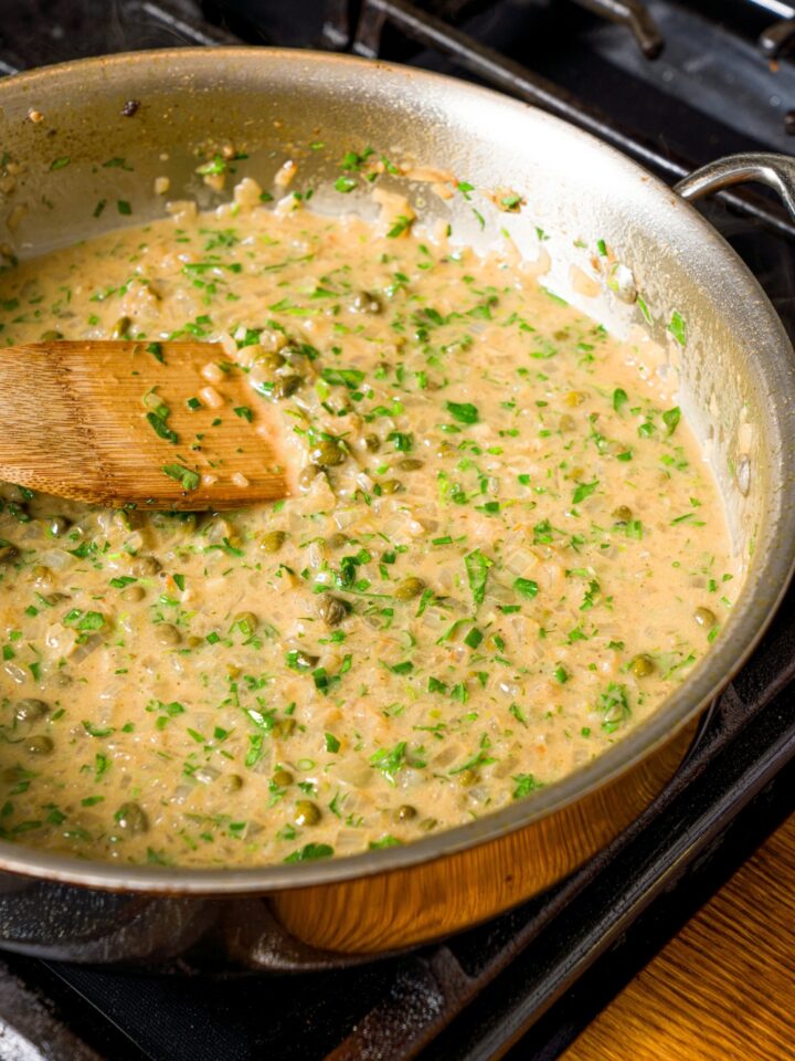 A skillet with chicken piccata sauce. The skillet is on a stove with a spoon stirring the mixture.