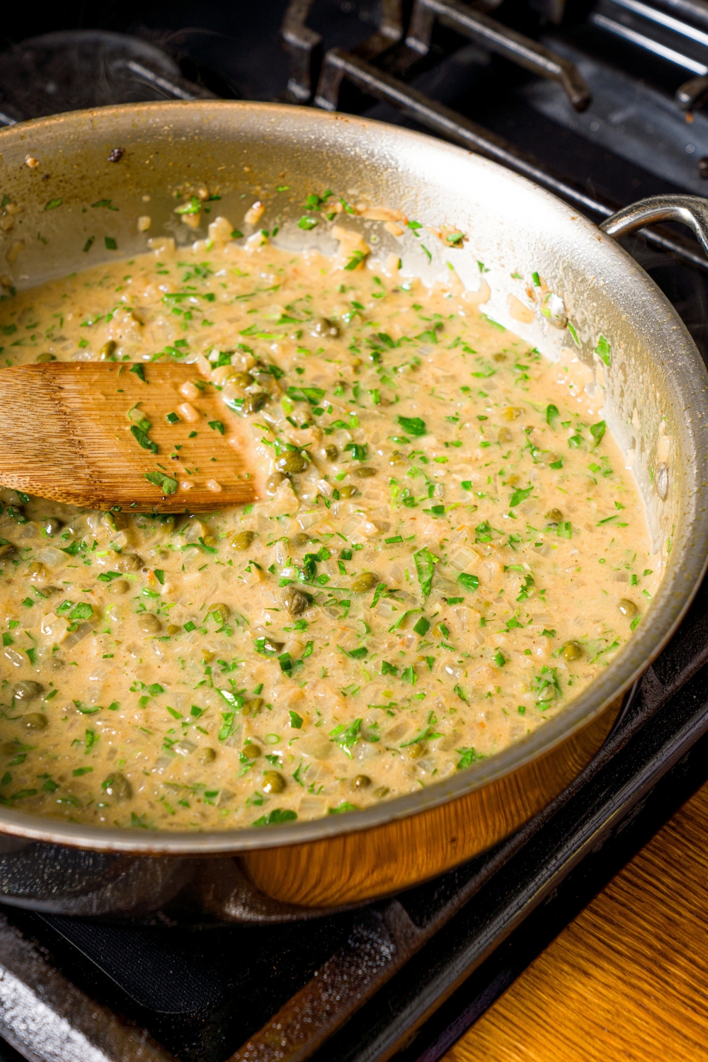 A skillet with chicken piccata sauce. The skillet is on a stove with a spoon stirring the mixture.