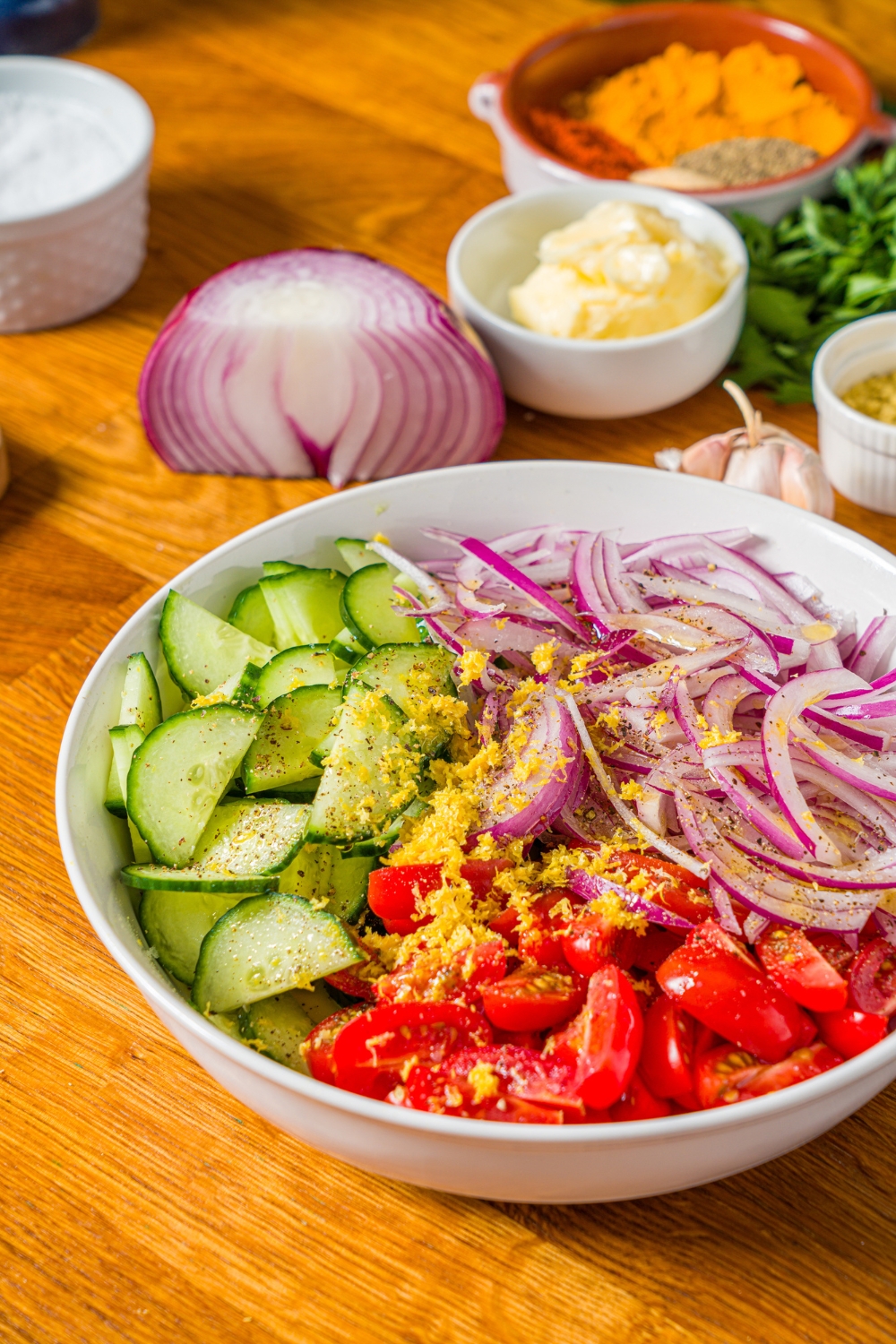 A white bowl with onion salad including sliced cucumbers, tomatoes, and red onion with lemon zest and olive oil. The bowl is on a wooden counter with other bowls of ingredients.