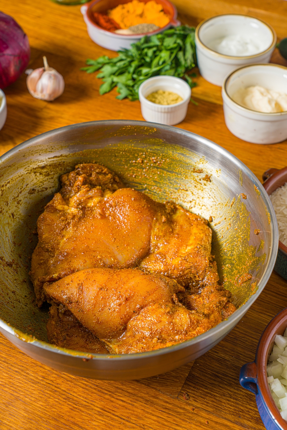 A large bowl with chicken thighs marinading in seasonings and oil. The bowl is on a wooden counter with additional bowls of ingredients.
