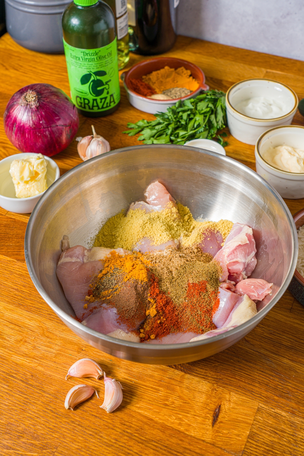 A large bowl with chicken thighs topped with seasonings including paprika, cumin, onion powder, turmeric, salt, and pepper. The bowl is on a wooden counter with bowls of ingredients.
