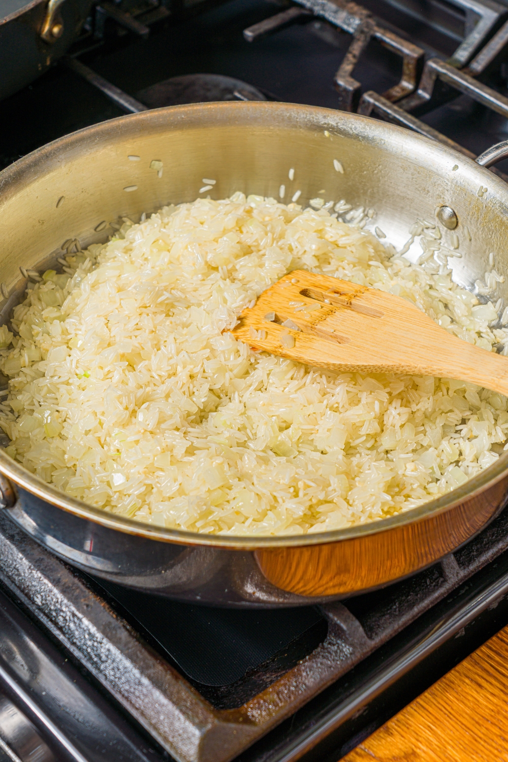 A skillet with diced onion, garlic, and rice cooking in butter. A wooden spoon is mixing the rice.