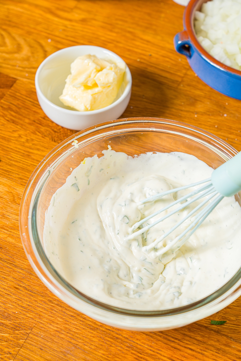 A glass bowl with a whisk mixing garlic sauce. The bowl is on a wooden counter with a bowl of butter.