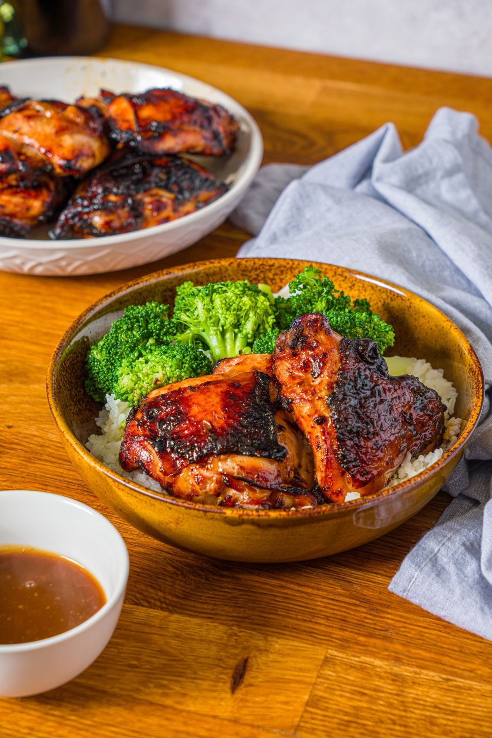 A bowl with chicken teriyaki served with broccoli and rice. The bowl is on a wooden counter with a bowl of cooked chicken and small bowl of teriyaki sauce.