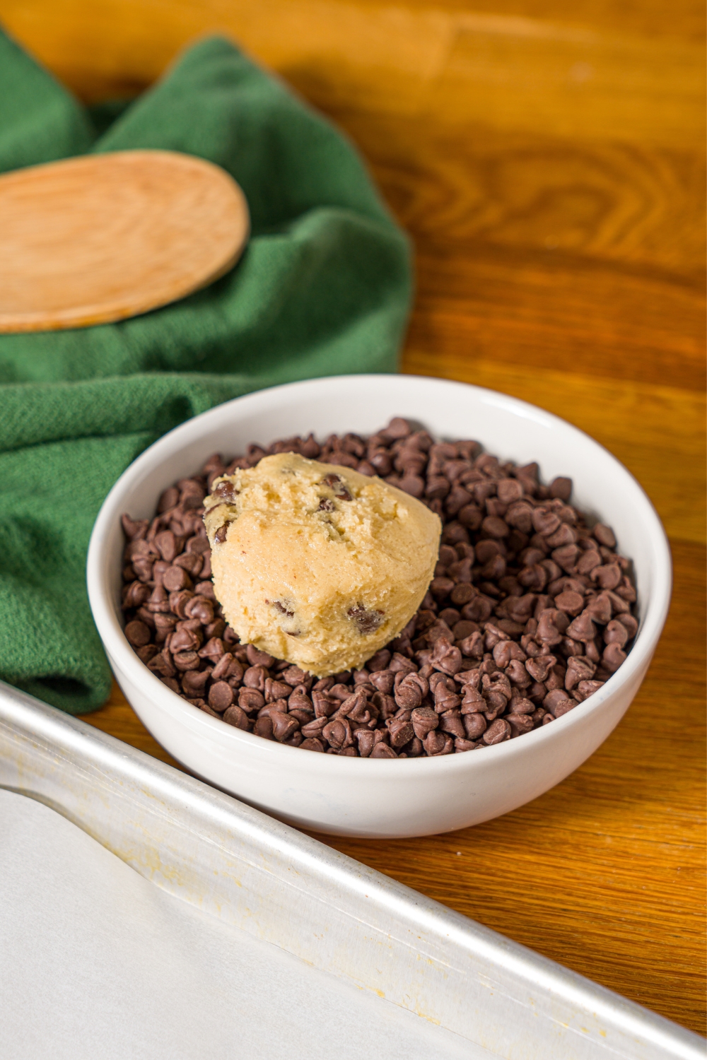 A chocolate chip cookie dough ball in a bowl of mini chocolate chips. The bowl is on a wooden counter with a green cloth napkin.