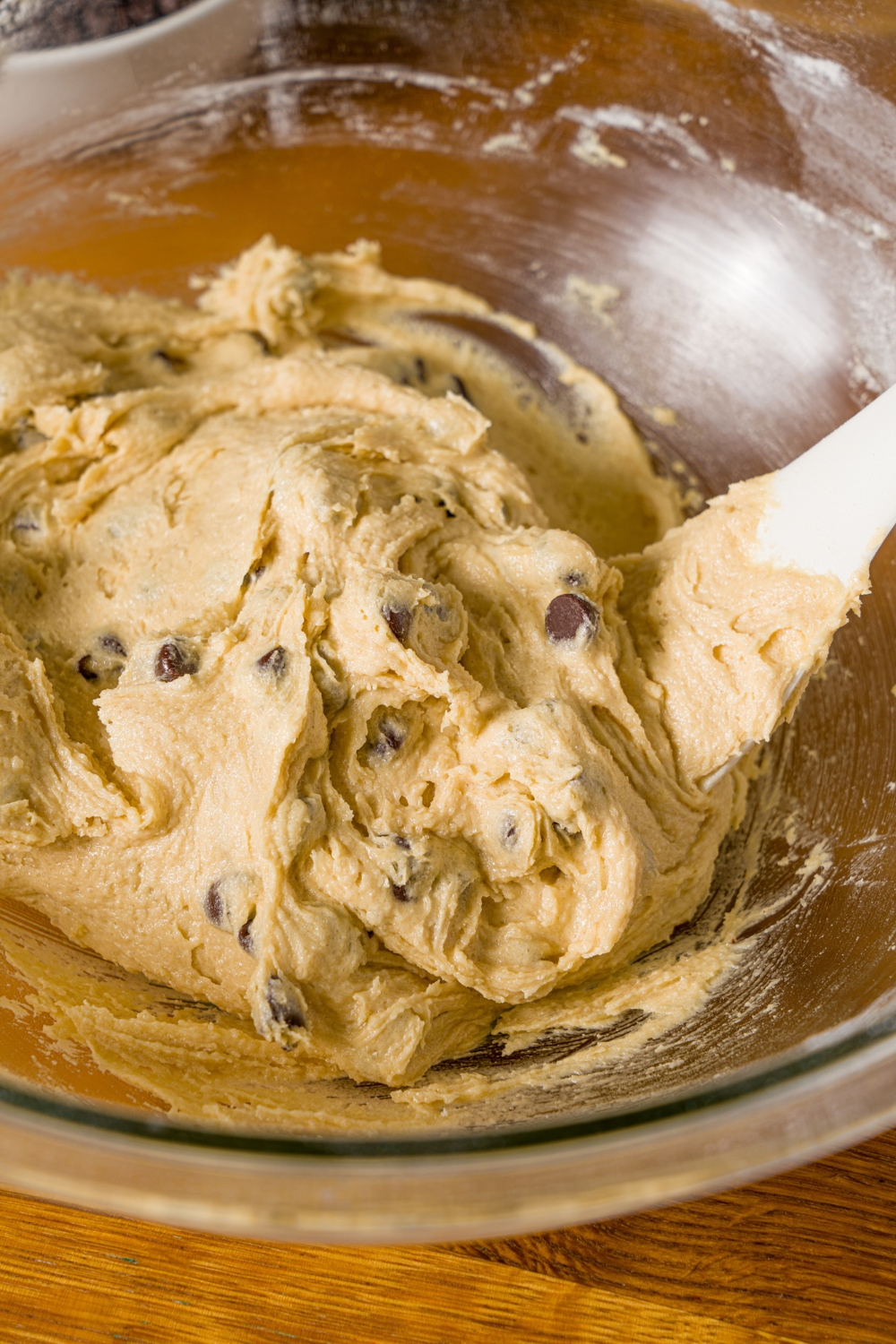 A glass bowl with a spatula mixing chocolate chip cookie dough without baking soda. The bowl is on a wooden counter.