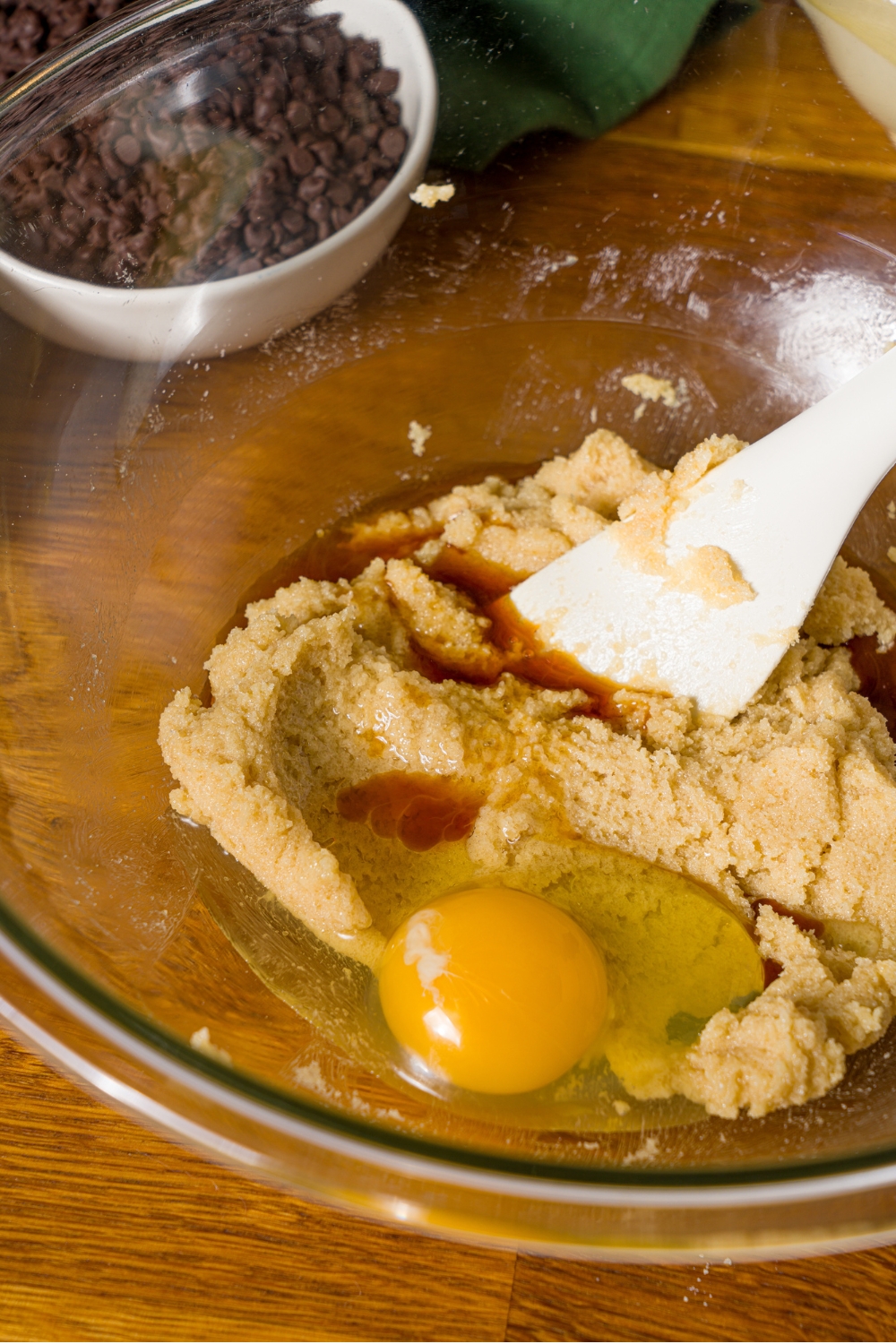 A glass bowl with spatula mixing creamed butter and brown sugar mixture, egg, and vanilla. The bowl is on a wooden counter with a bowl of chocolate chips.
