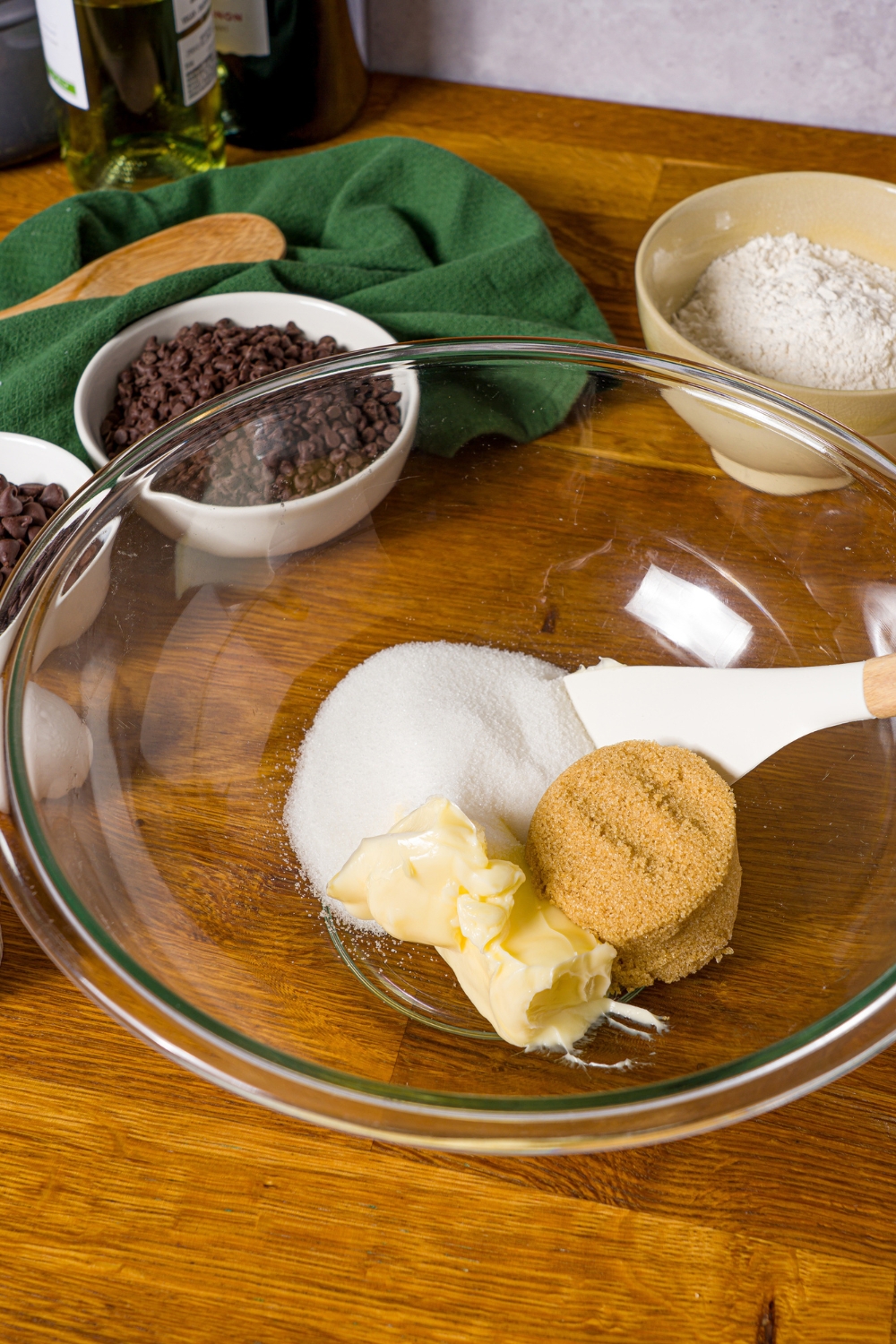 A glass bowl with a spatula mixing sugar, brown sugar, and butter. The bowl is on a wooden counter with additional bowls of ingredients and a green cloth napkin.
