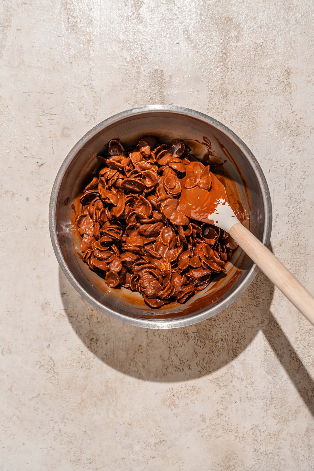 A mixing bowl with a wooden spoon mixing chocolate cornflake crunch mixture. The bowl is on a tan counter.