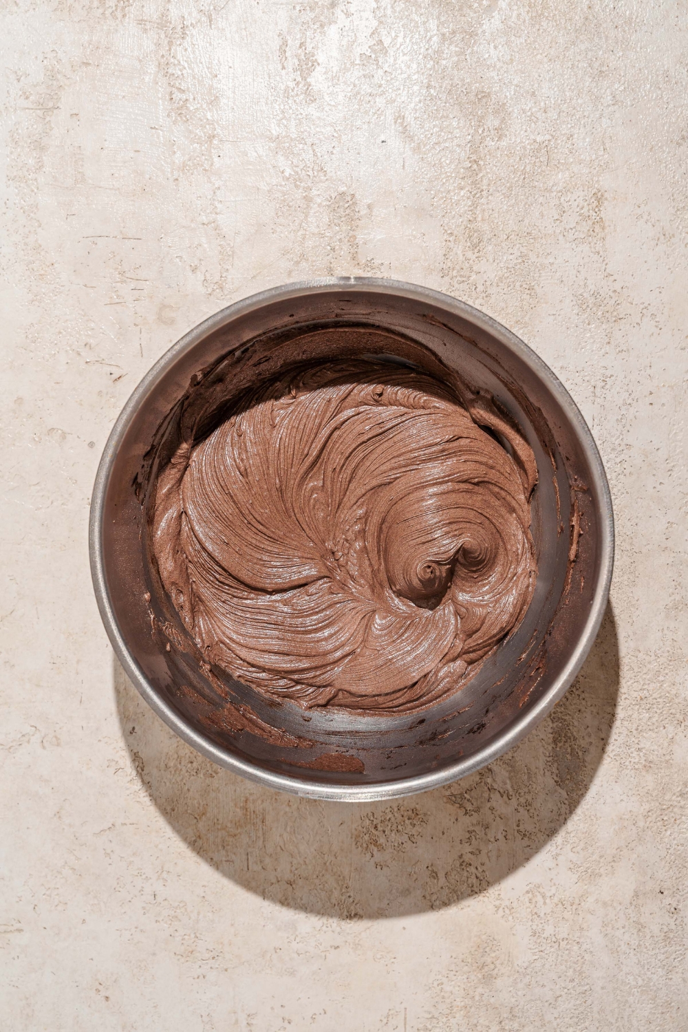 A large mixing bowl whipped chocolate cream cheese frosting. The bowl is on a tan counter.