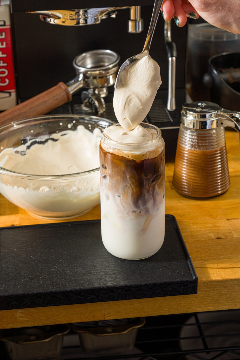 A tall glass with iced cookie butter latte. A spoon is adding cookie butter cold foam on top. The glass is on a wooden counter with a bowl of cold foam and an espresso machine.