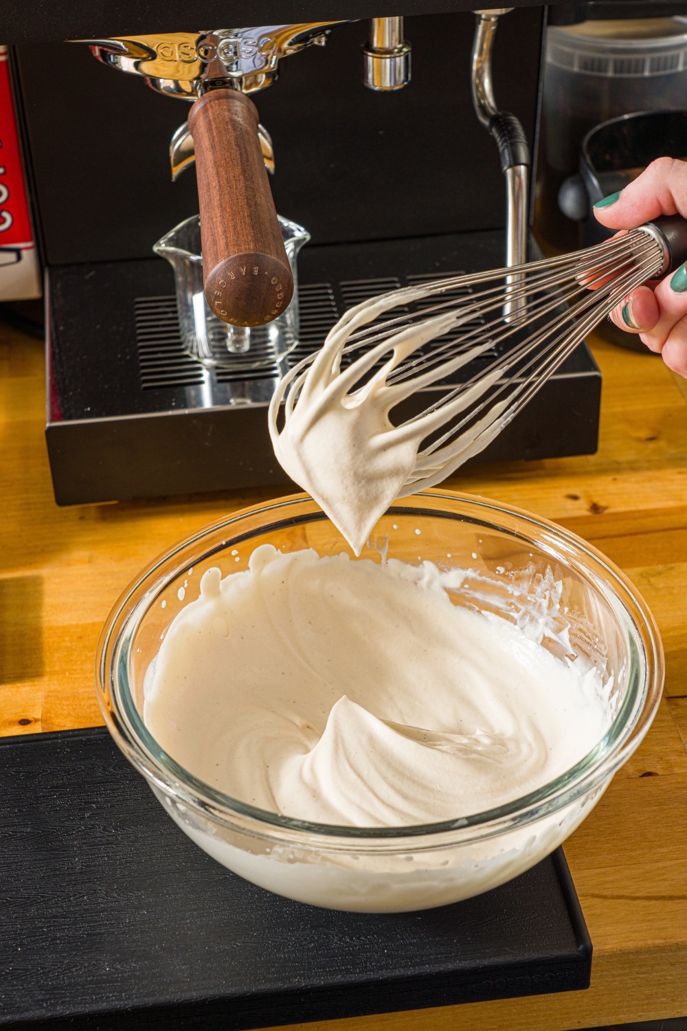 A glass bowl with a whisk mixing cookie butter cold foam. The bowl is on a wooden counter with an espresso machine.