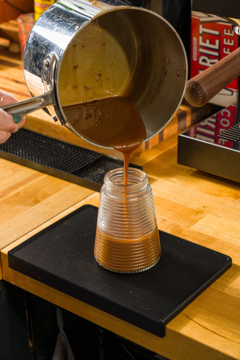 A small jar with a saucepan of cookie butter syrup being poured into the jar. The jar is on a wooden counter.