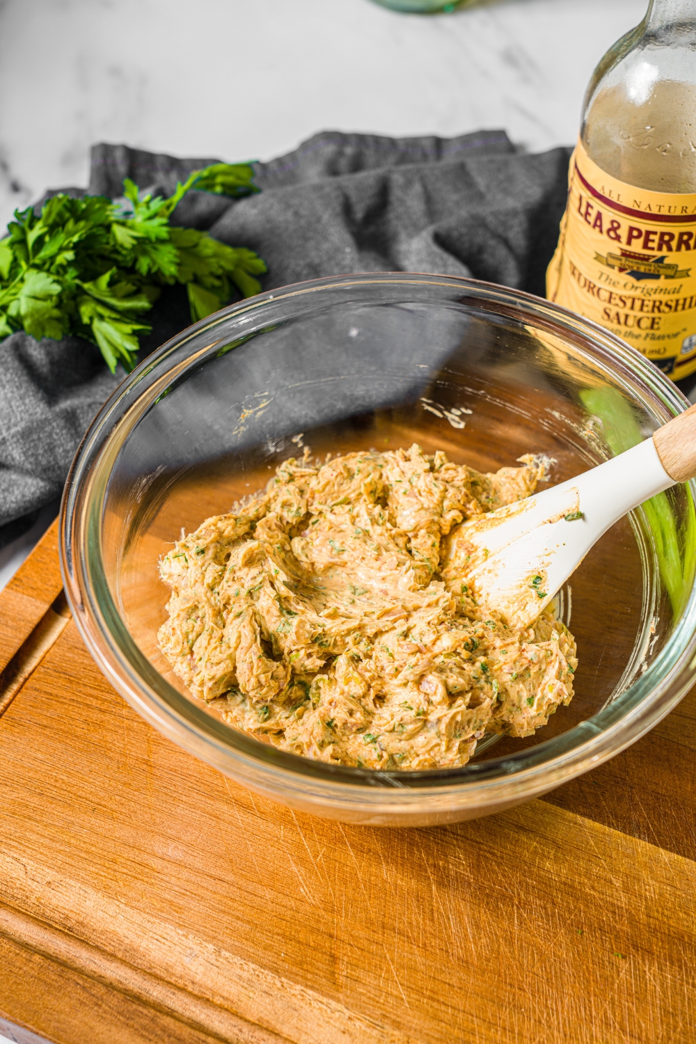 A glass bowl with a spatula mixing ingredients to make cowboy butter including butter, mustard, Worcestershire sauce, and seasonings. The bowl is on a wooden board with a grey cloth napkin.