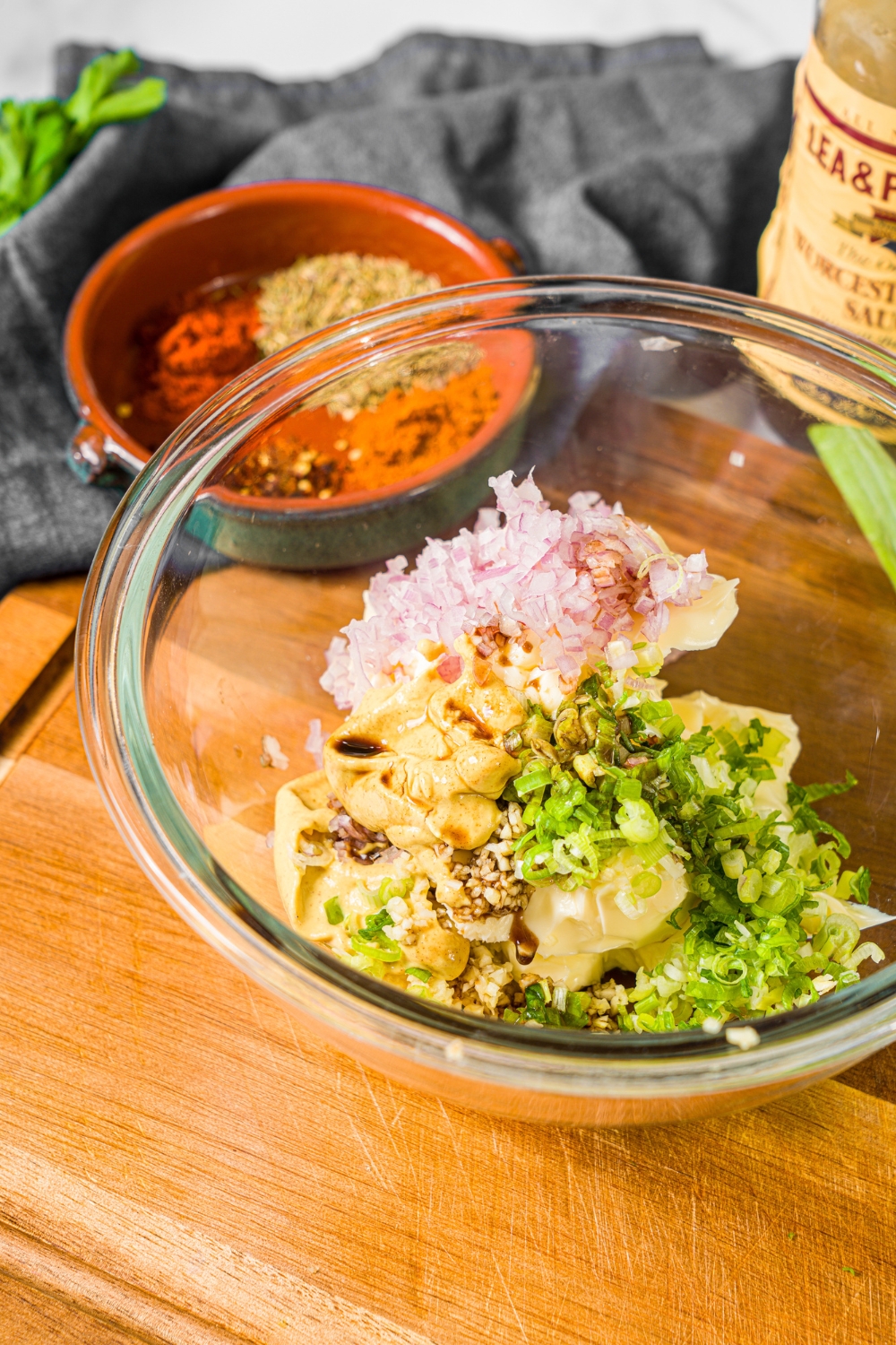 A glass bowl with ingredients to make cowboy butter including butter, sliced green onion, sliced red onion, mustard, and Worcestersire sauce. The bowl is on a wooden counter with a small bowl of seasonings.